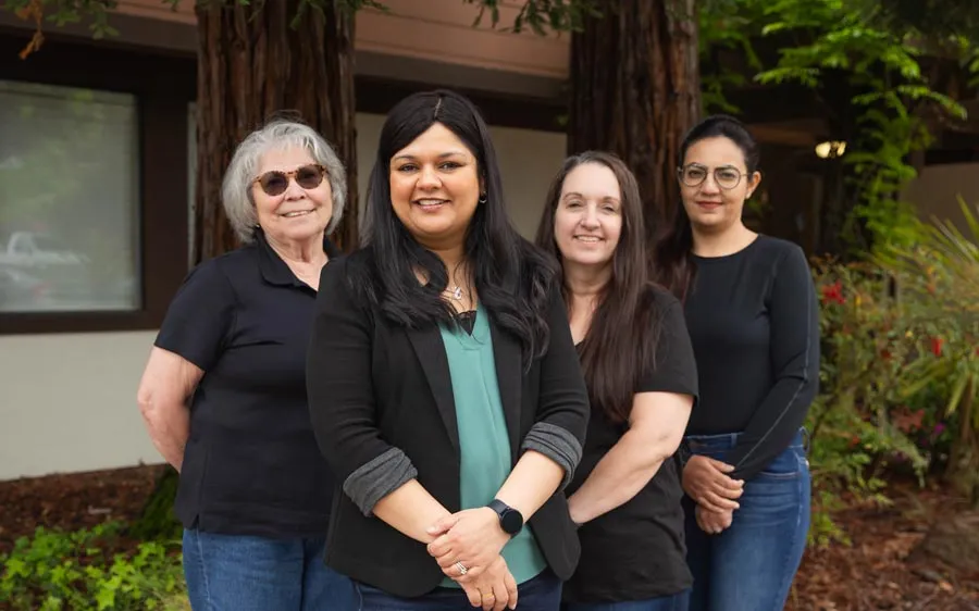 Group of four diverse women standing outdoors smiling in casual and professional attire near trees and greenery