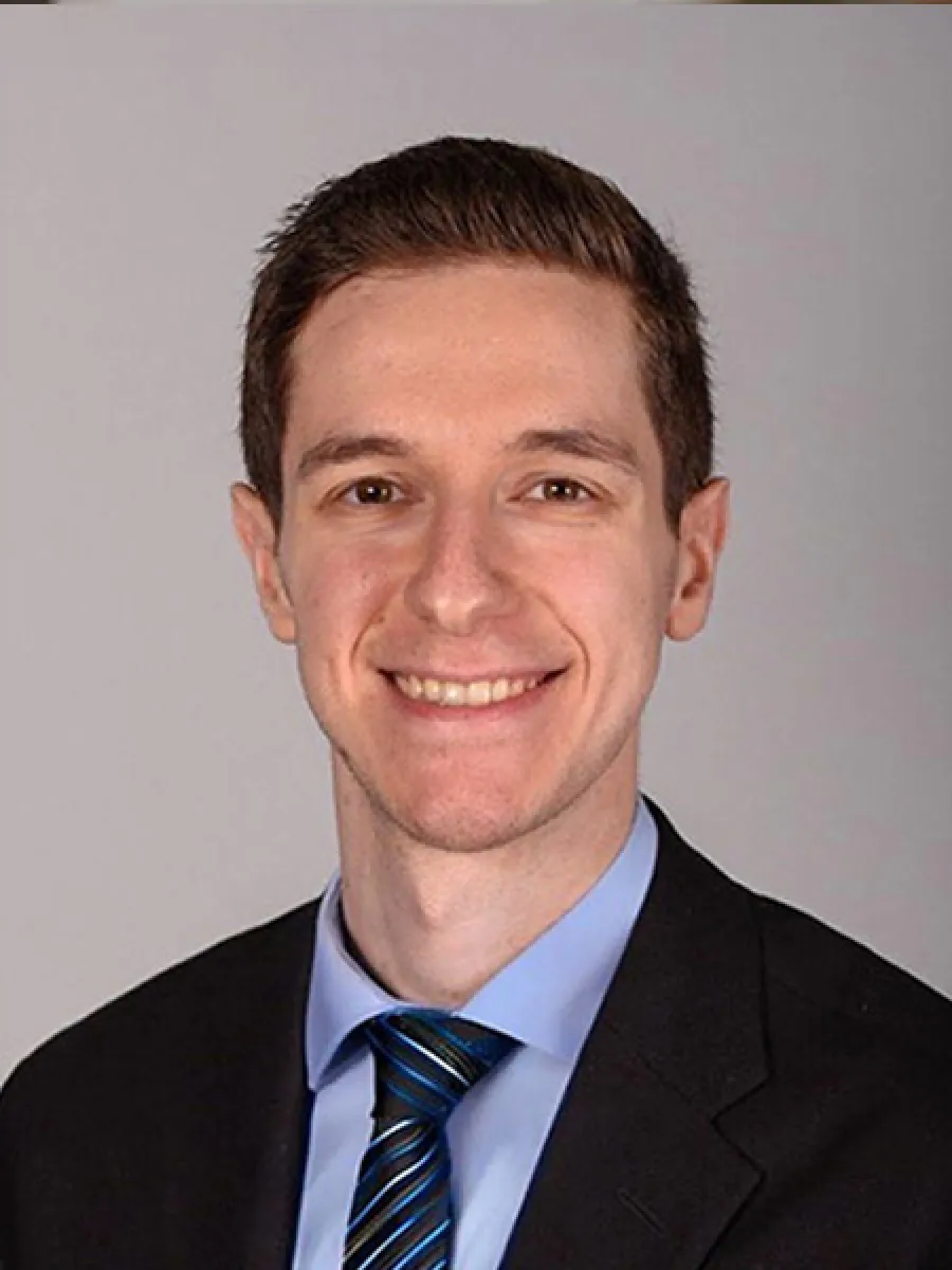 Professional headshot of a young man smiling, wearing a black suit, blue shirt, and striped tie against a gray background.