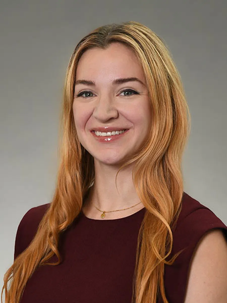 Smiling woman with long strawberry blonde hair wearing a maroon sleeveless top against a gray background