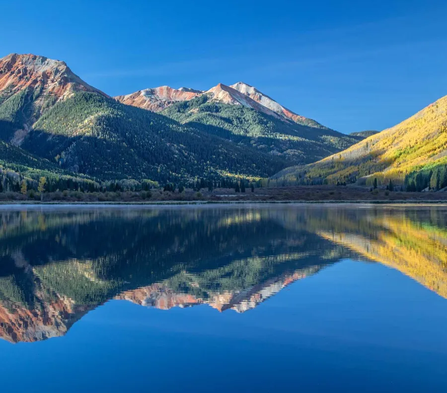 Mountain range with red and green slopes reflected clearly in calm lake under a bright blue sky