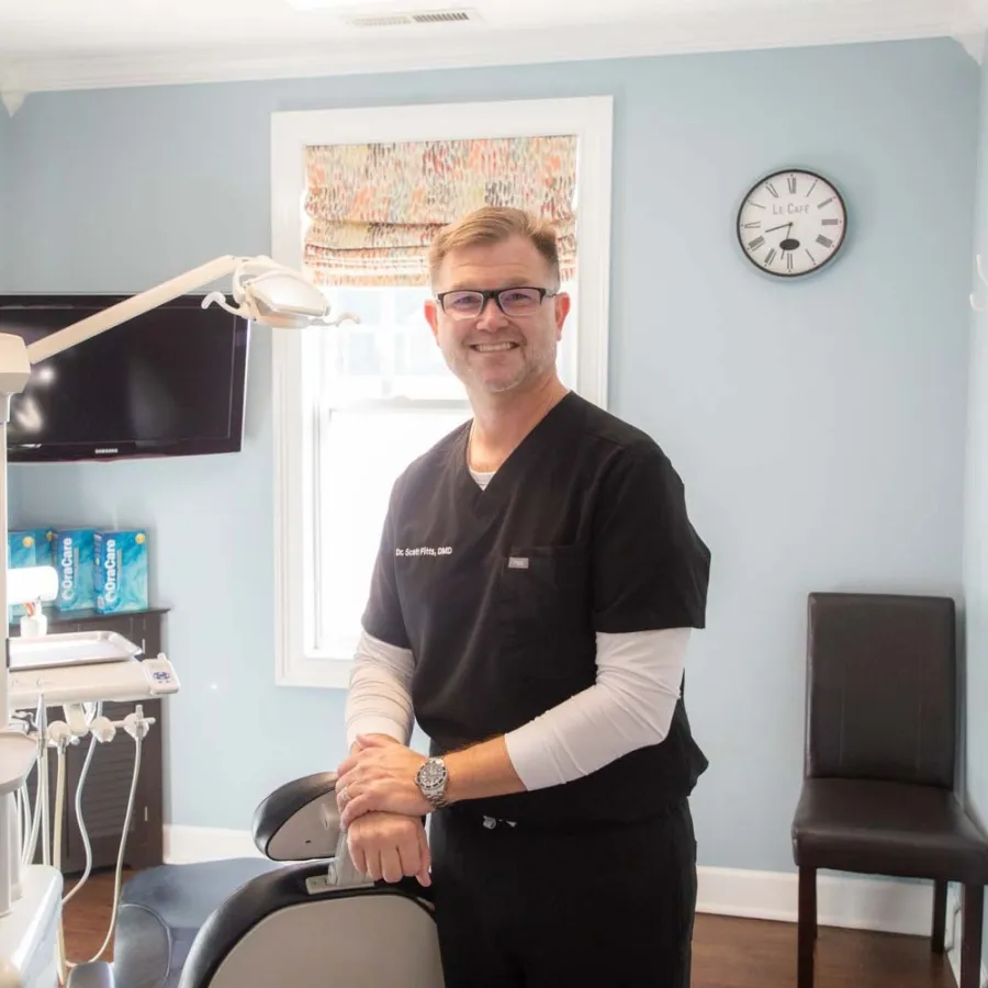 Smiling male dentist in black scrubs standing in a bright dental office next to dental chair and equipment.
