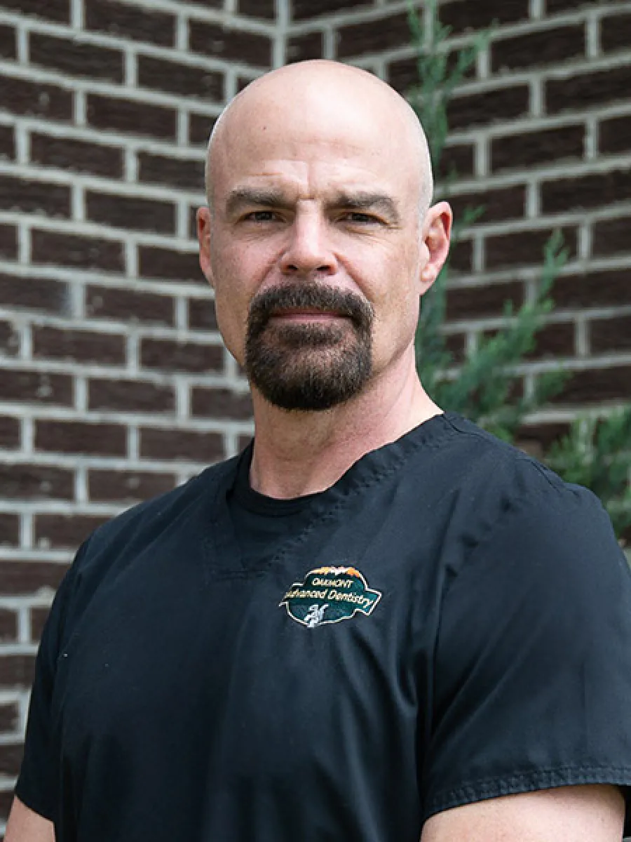 Portrait of a bald man with a goatee wearing a black Oremont Advanced Dentistry uniform against a brick wall