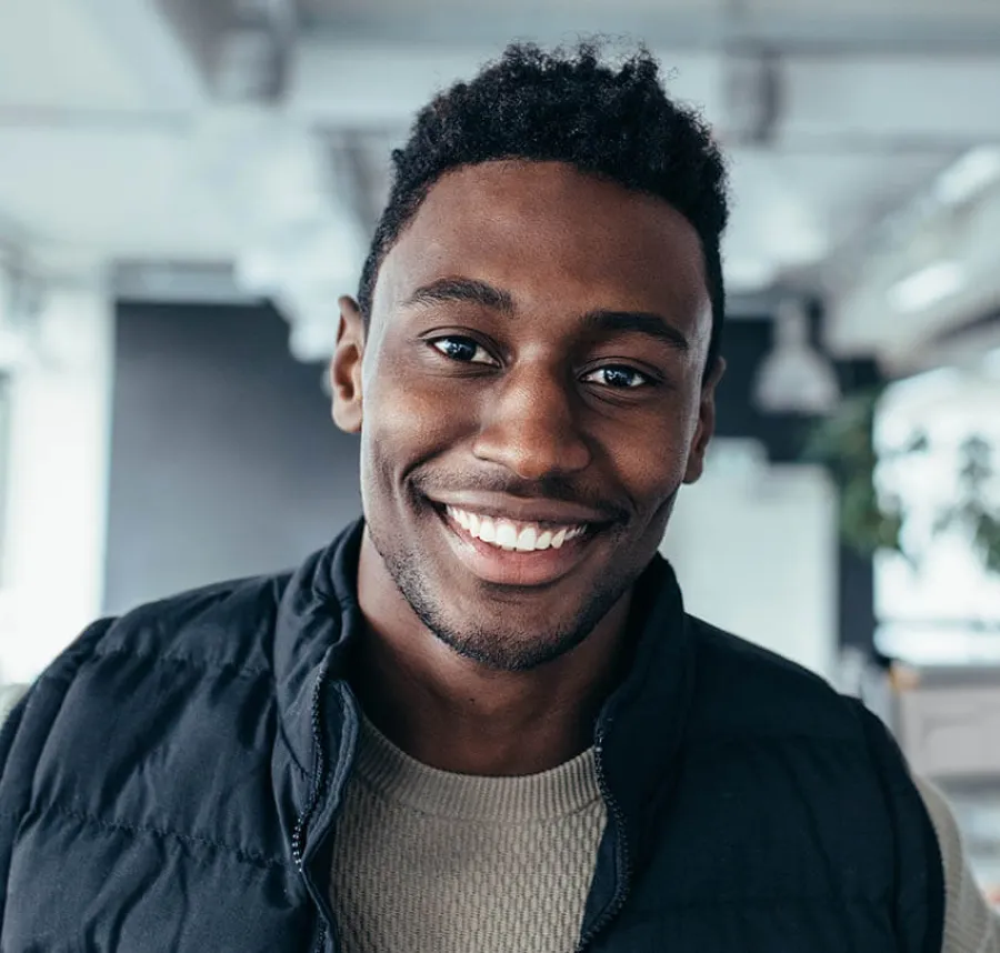 Smiling young African American man wearing a black jacket in a modern office setting with blurred background.