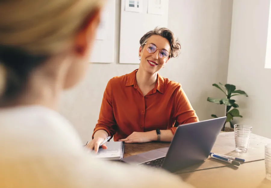 Woman in orange shirt smiling and talking while working on laptop during a meeting in bright office.