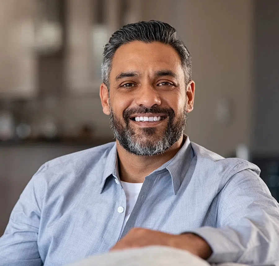 Smiling middle-aged man with salt and pepper beard wearing a light blue shirt in a cozy indoor setting.
