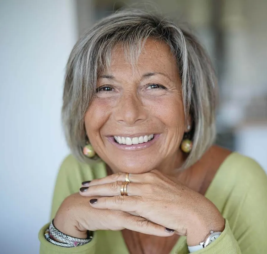 Smiling middle-aged woman with gray hair resting her chin on hands, wearing green top and jewelry indoors.