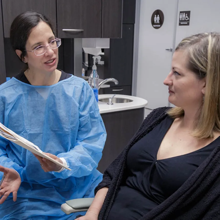 Healthcare professional in blue gown consulting a seated woman in a medical office with sink and restroom signs.