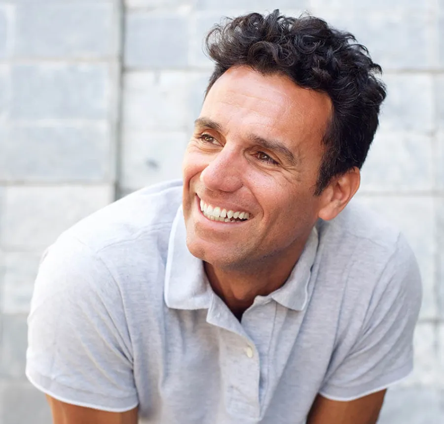 Smiling man with curly dark hair wearing a light gray polo shirt against a stone wall background.