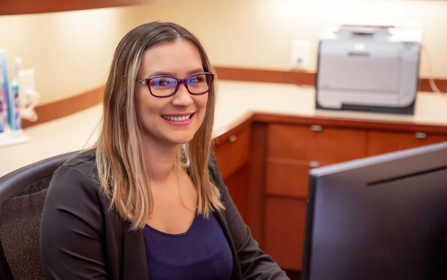 Smiling woman with glasses sitting at office desk working on a computer in a professional workspace.