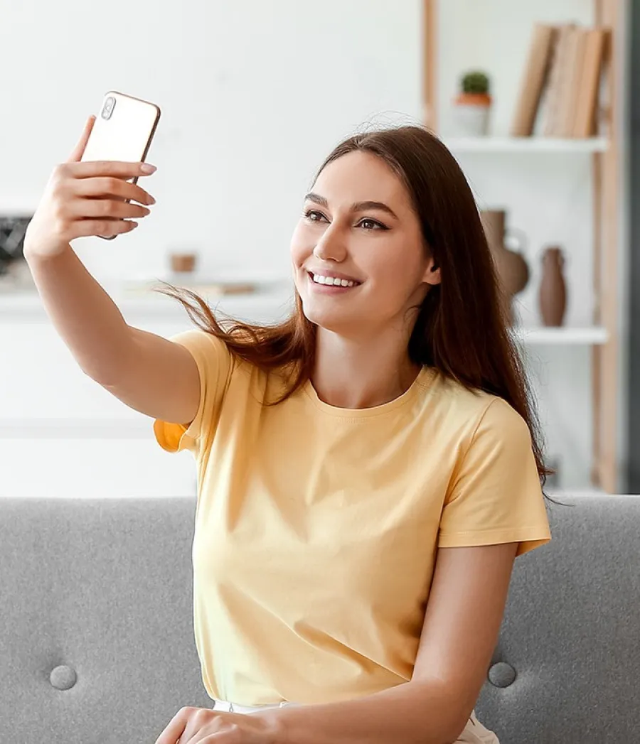 Smiling young woman in yellow shirt sitting on a couch taking a selfie with her smartphone indoors