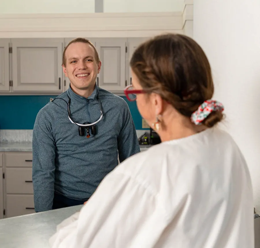 Dentist with magnifying glasses talking and smiling to a female patient in a clinic setting.