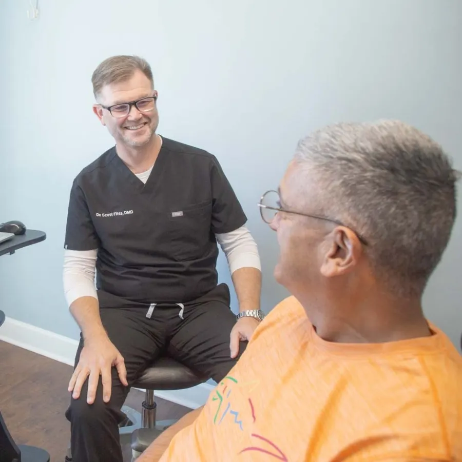 Dentist consulting a male patient in a bright office with dental 3D model on laptop screen.