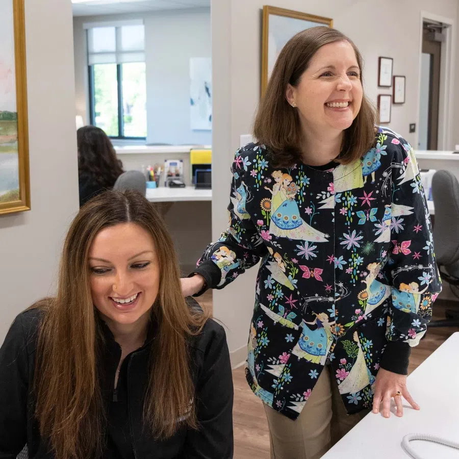 Two women smiling and interacting in a bright office with desks and computers in the background