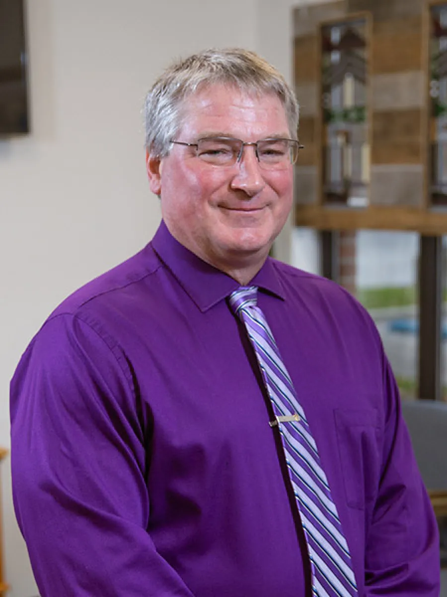 Smiling middle-aged man wearing a purple dress shirt and striped tie with glasses indoors.