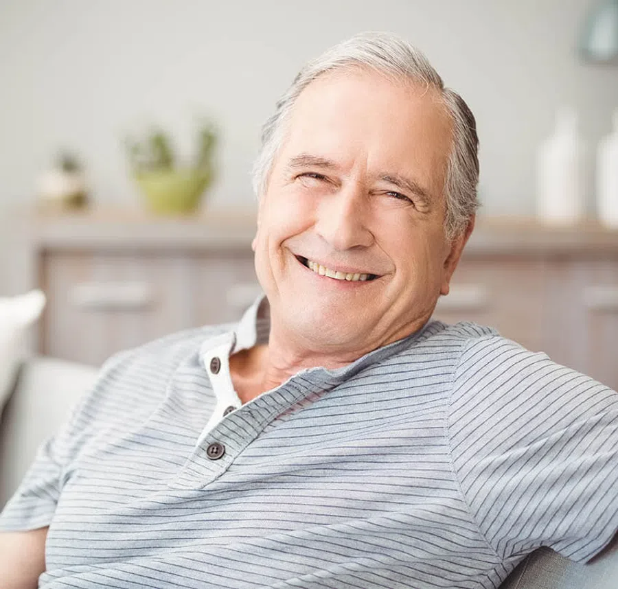 Smiling elderly man in a striped shirt sitting comfortably on a couch indoors with soft background.