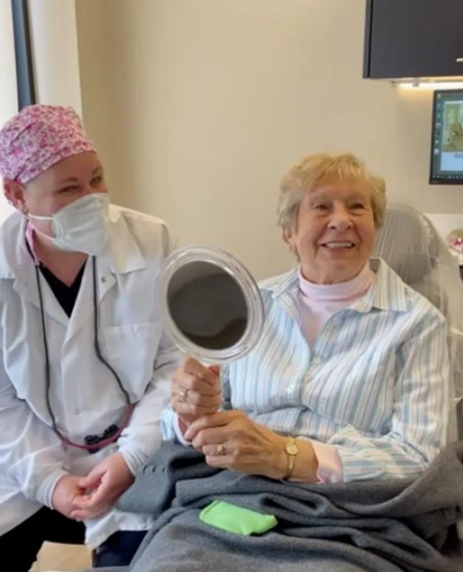 Smiling elderly woman holding a mirror next to a masked healthcare worker in a medical setting.