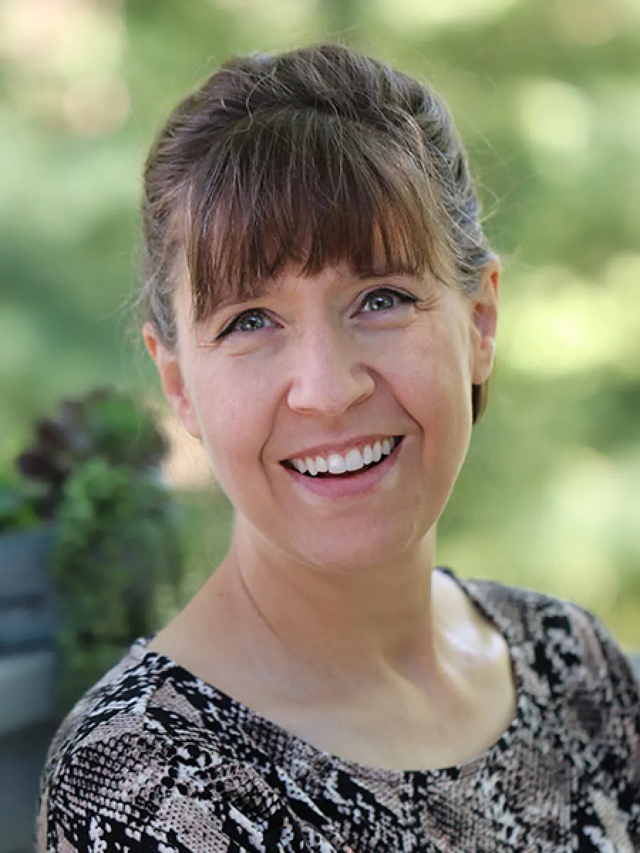 Smiling woman with brown hair and bangs wearing a patterned top outdoors with blurred greenery background