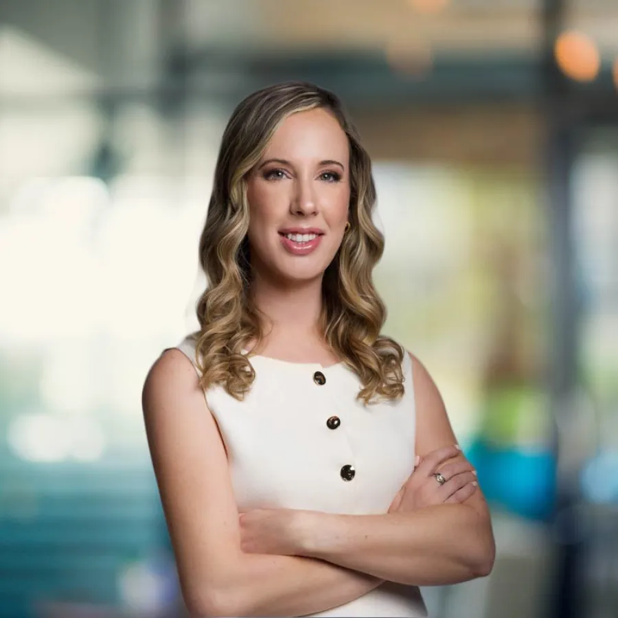 Confident woman with curled blonde hair wearing a white sleeveless dress posing with arms crossed indoors.