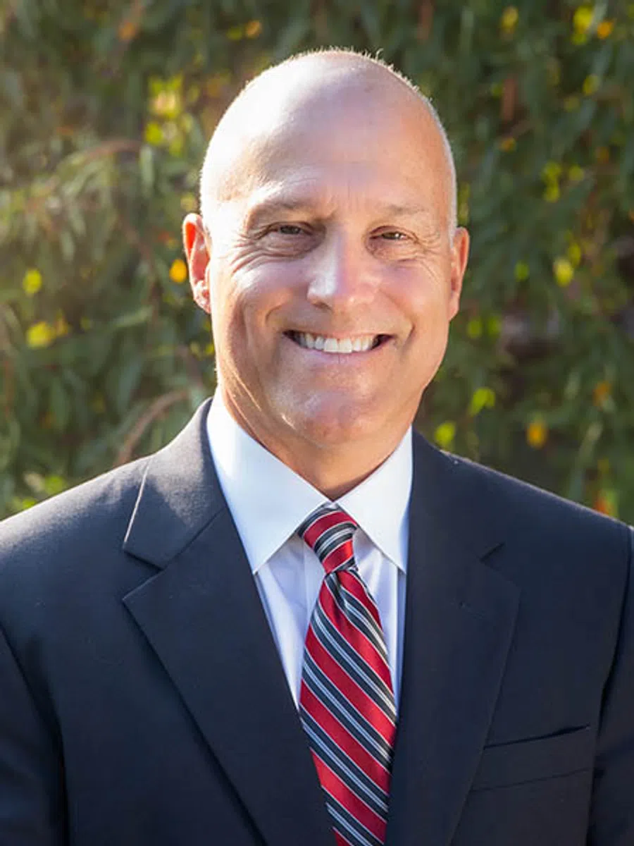 Smiling bald man in a dark suit and red striped tie posing outdoors with blurred green foliage background