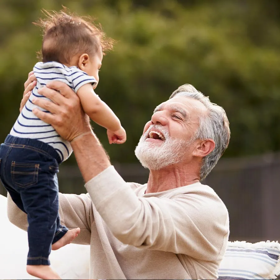 A joyful grandfather lifts his smiling baby grandson in a sunny outdoor setting.