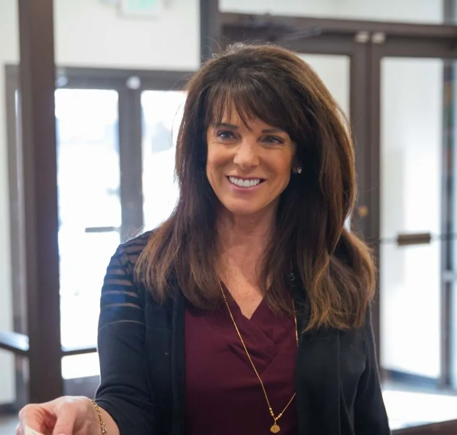 Smiling woman handing a card to another person in a bright indoor setting with large windows and doors.