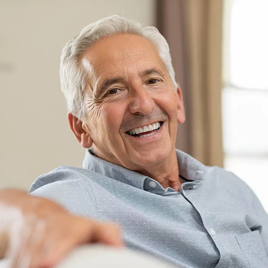 Smiling elderly man with white hair wearing a light blue shirt sitting comfortably indoors.