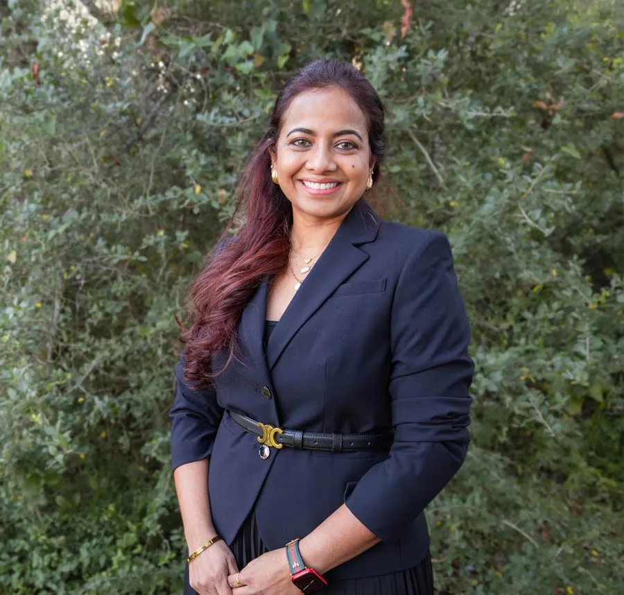 Smiling woman in a black blazer standing outdoors with green foliage background, wearing jewelry and a smartwatch.