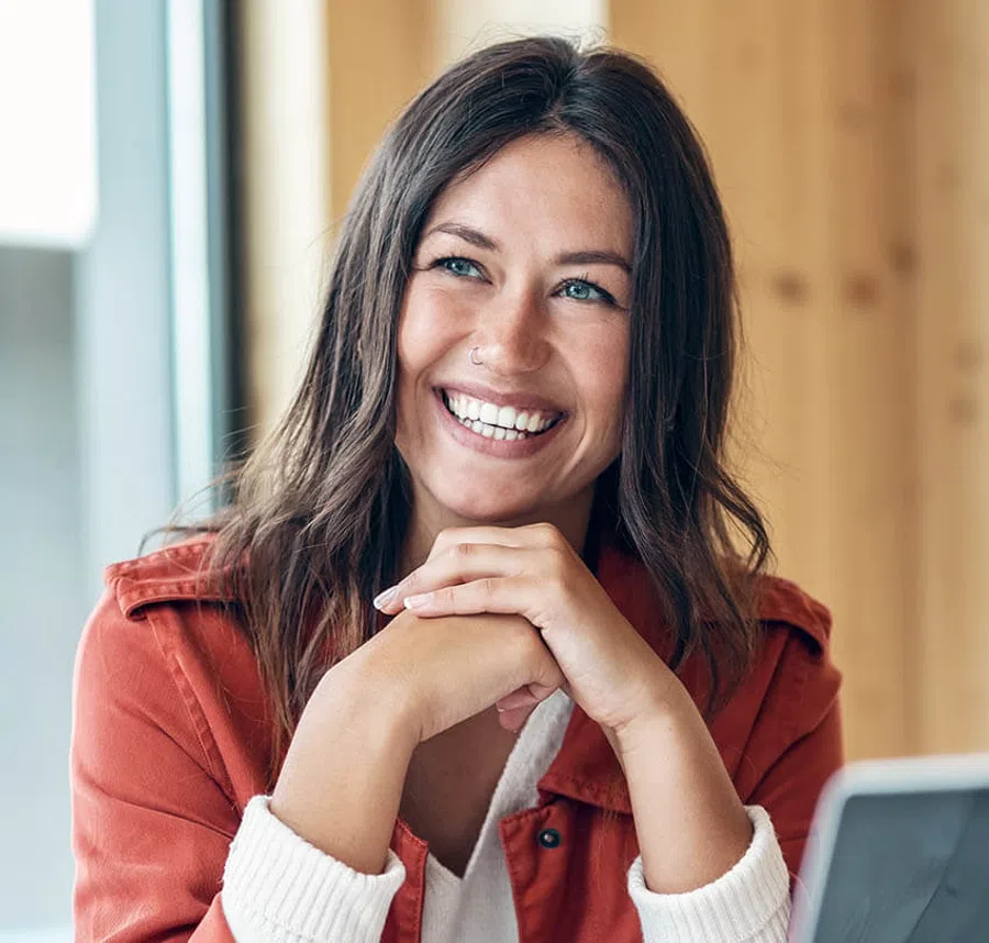Smiling woman with long dark hair wearing a rust-colored jacket and white sweater, sitting indoors by a window.