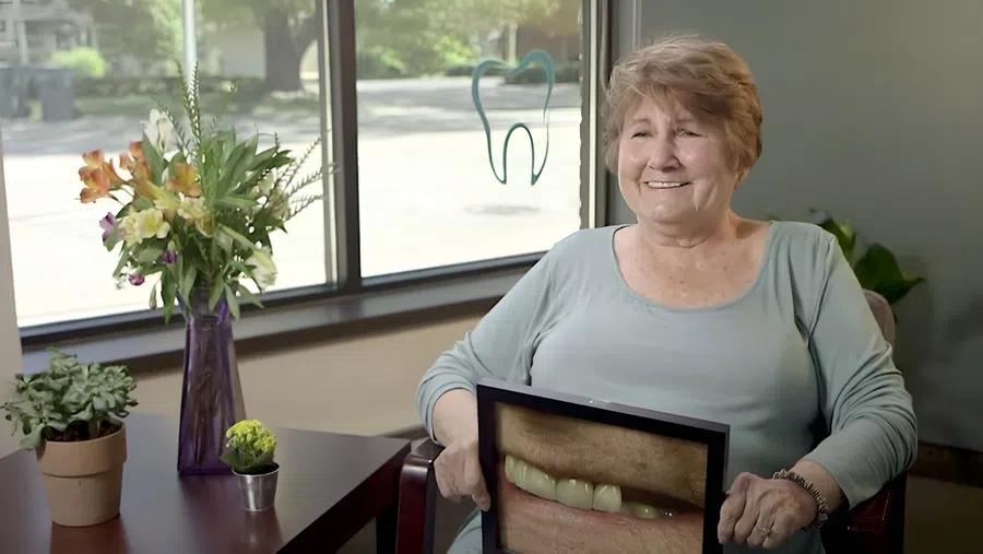 Smiling elderly woman holding framed photo of dental implants in a bright dental clinic waiting room.