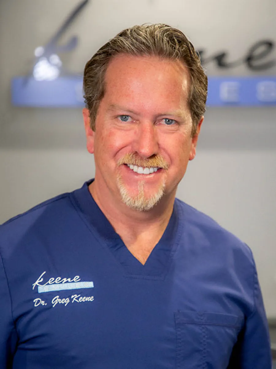Smiling male doctor in blue scrubs with Keene Dental embroidered, standing in a dental office.
