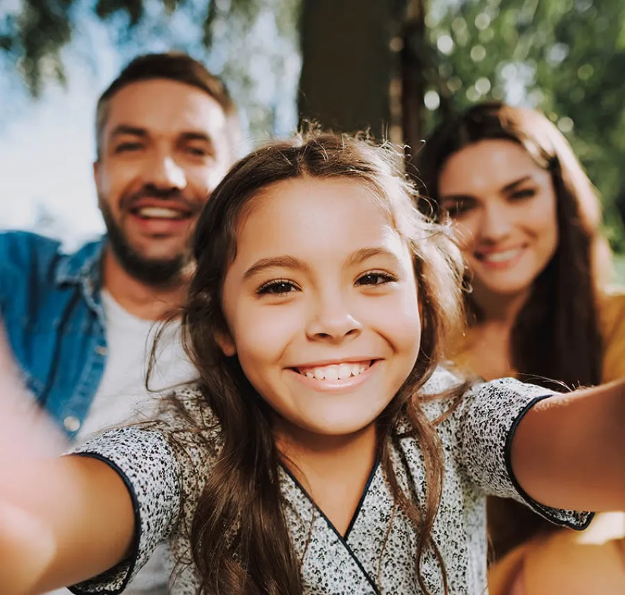 Smiling young girl taking selfie with her happy parents outdoors on a sunny day.