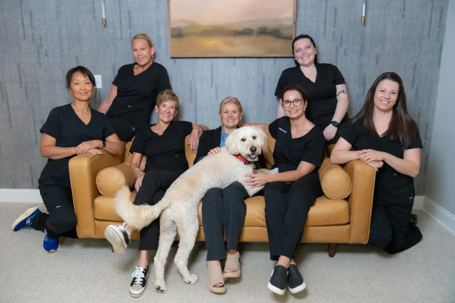Group of seven women in black uniforms posing with a large white dog on a tan couch in a cozy room.