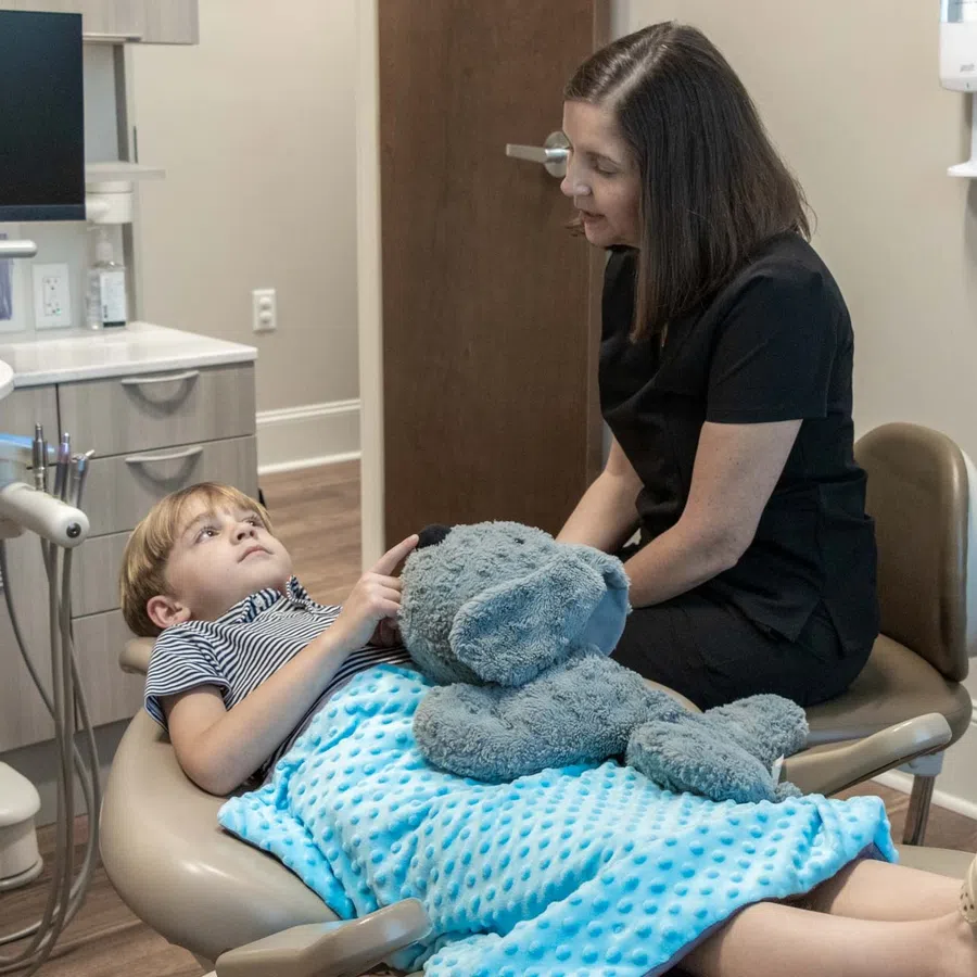 Child patient lying on dental chair with blue blanket and stuffed elephant toy, talking to female dentist in black scrubs