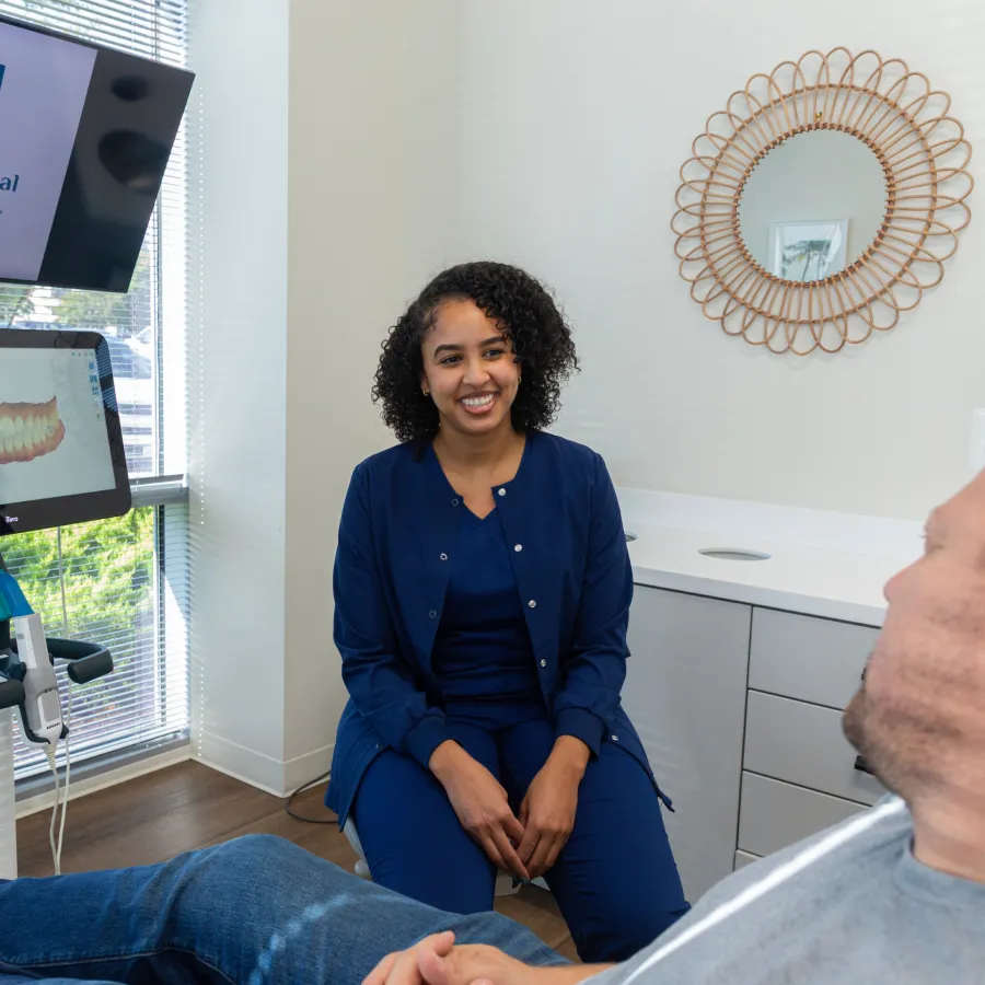 Smiling female dental professional in navy scrubs consulting with a male patient in a modern dental office.