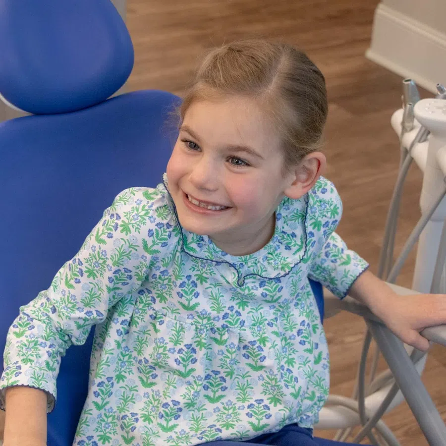 Smiling young girl in floral dress sitting in a blue dental chair at a dental clinic.