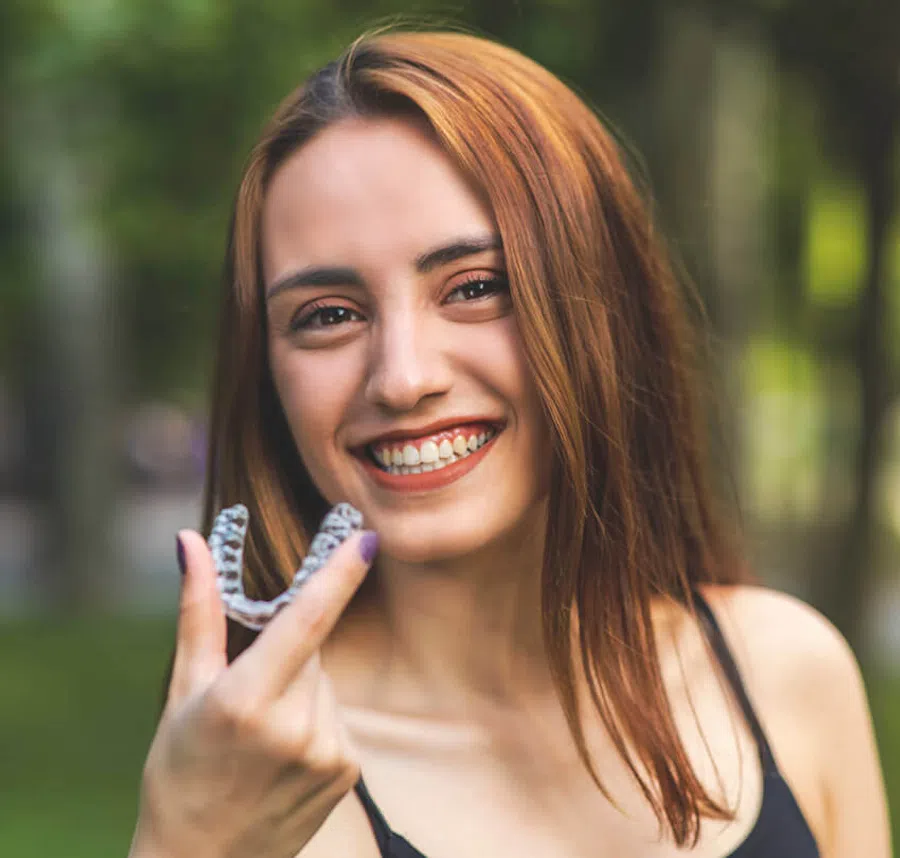 Smiling young woman holding a clear dental aligner outdoors in a green park setting