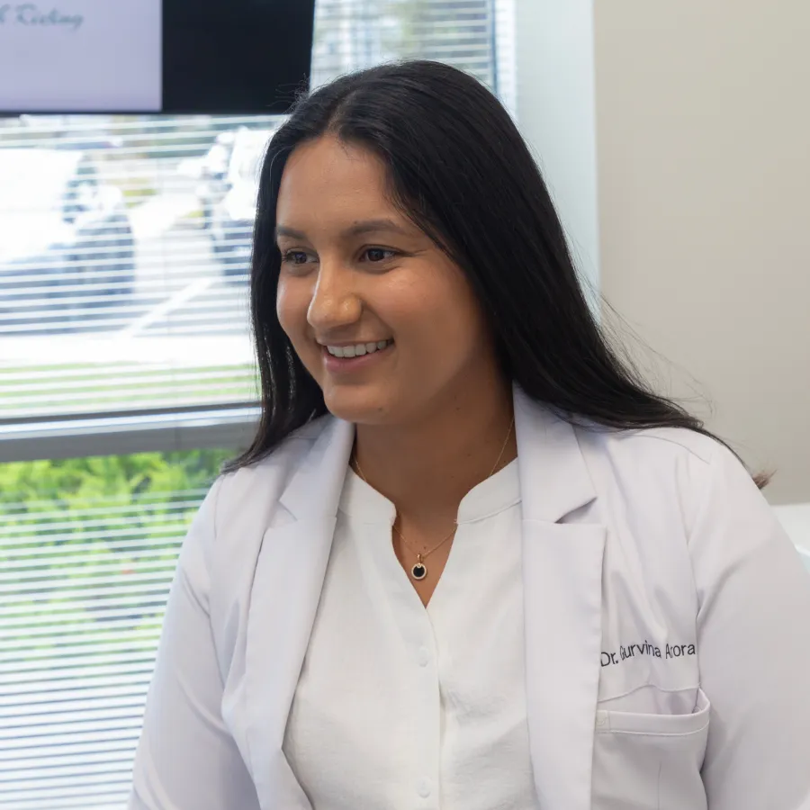 Smiling female doctor wearing a white coat in a bright office with window blinds and greenery outside.