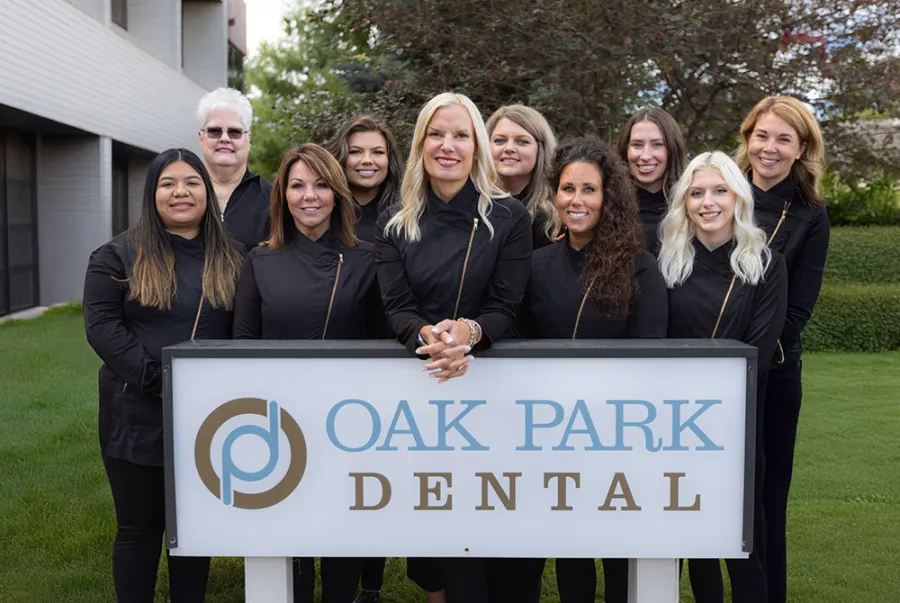 Dental team of nine women standing behind Oak Park Dental sign outside building with greenery.