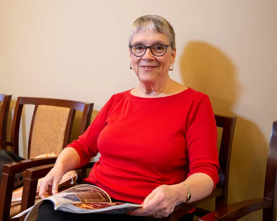 Smiling elderly woman in red shirt sitting on a chair reading a magazine in a waiting room with beige walls.