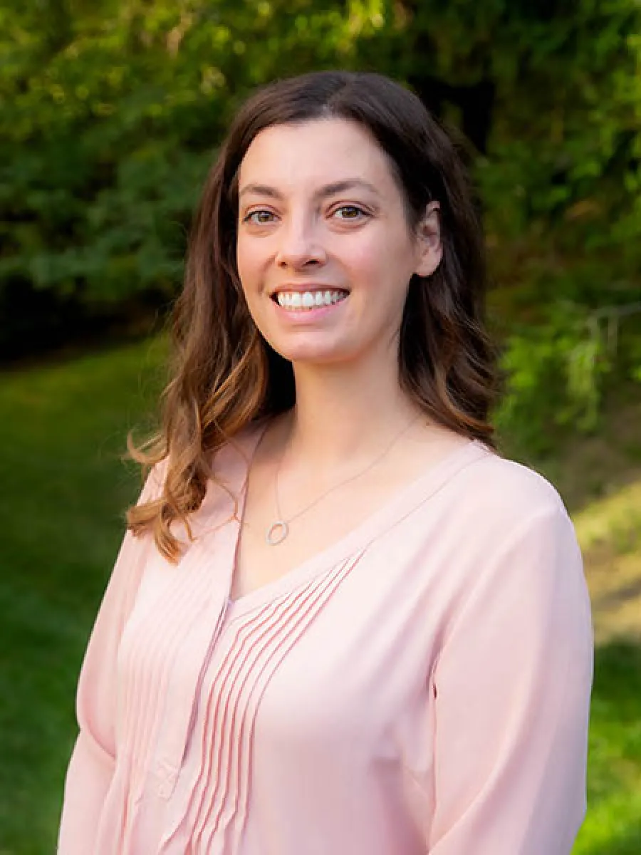 Smiling woman with brown hair wearing a light pink blouse standing outdoors with greenery in the background