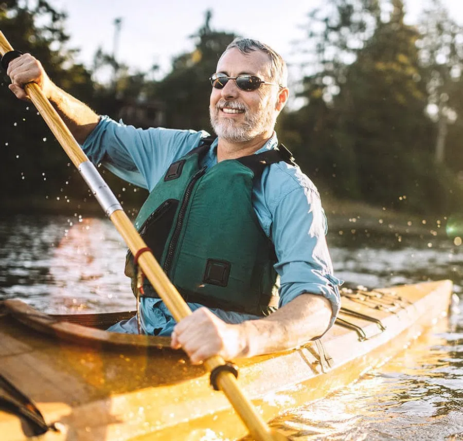 Smiling man wearing sunglasses and life jacket paddling a wooden kayak on a sunny lake with trees in the background
