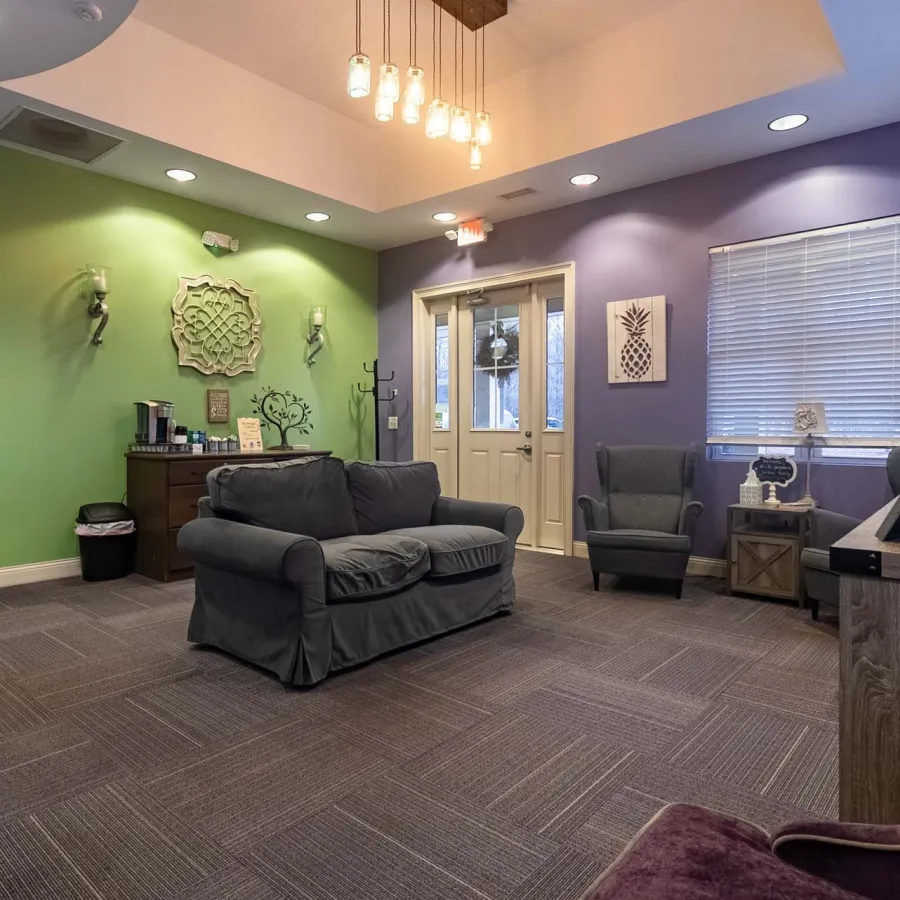 Cozy waiting room with green and purple walls, gray sofa, armchairs, decorative wall art, and soft lighting.