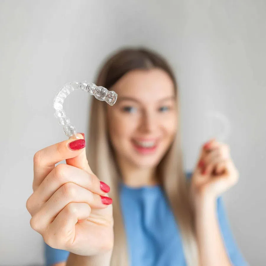 Smiling woman holding clear dental aligners for teeth straightening in a bright, neutral setting.