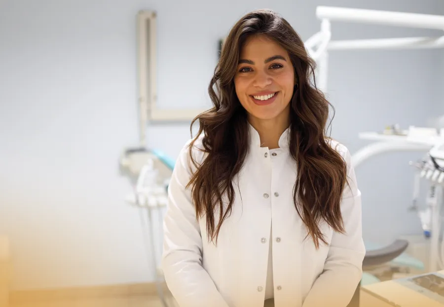 Smiling female dentist in white coat standing in a dental clinic with equipment in the background.