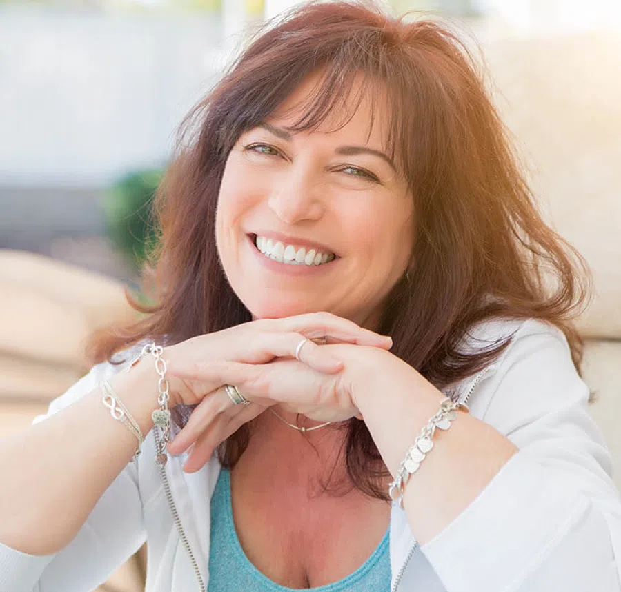 Smiling middle-aged woman with brown hair wearing a white jacket and turquoise top relaxing with hands clasped.