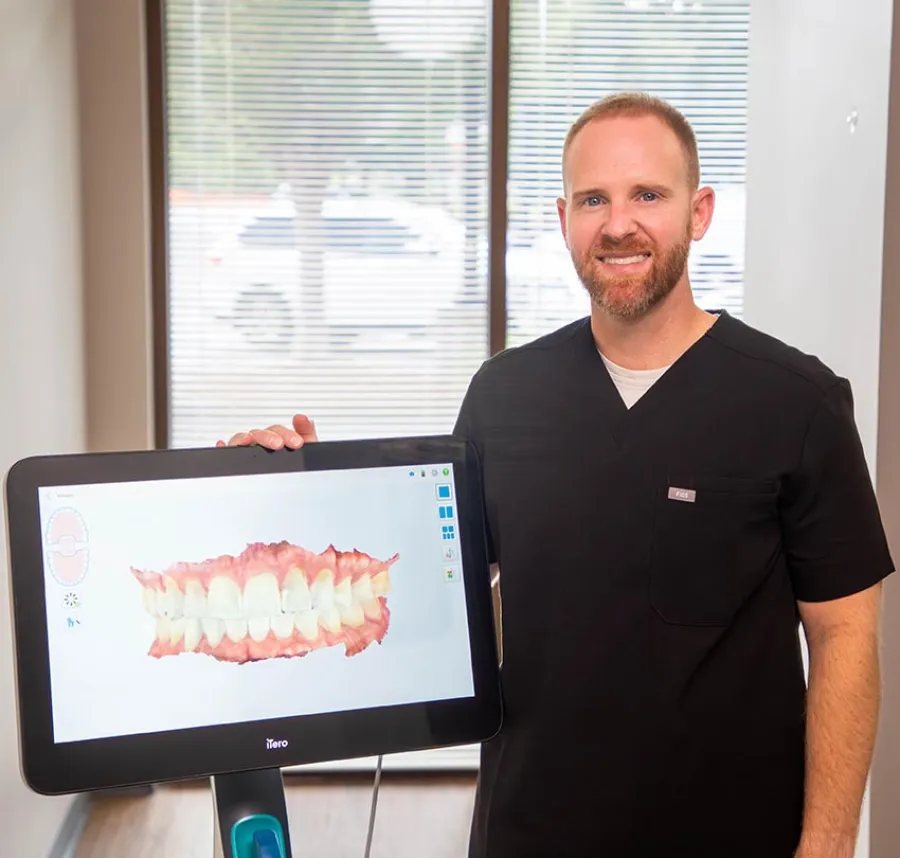 Male dentist in black scrubs displaying digital 3D dental scan on screen in bright office with window blinds.