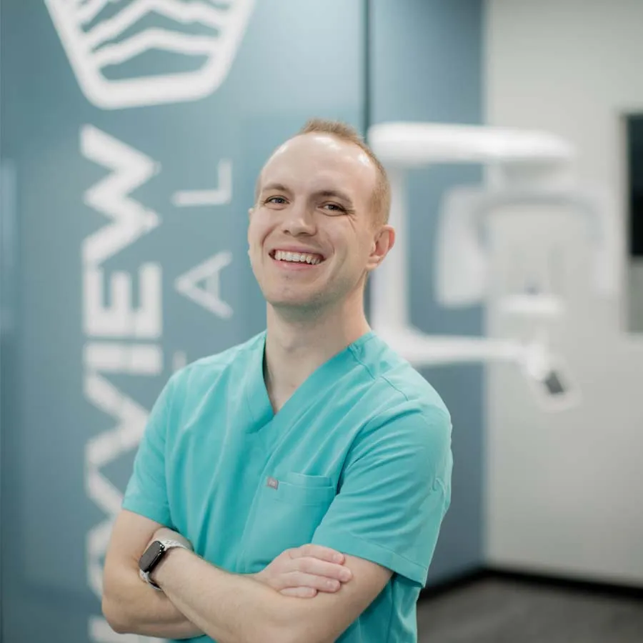 Smiling male healthcare professional in teal scrubs standing with arms crossed in a modern medical office.