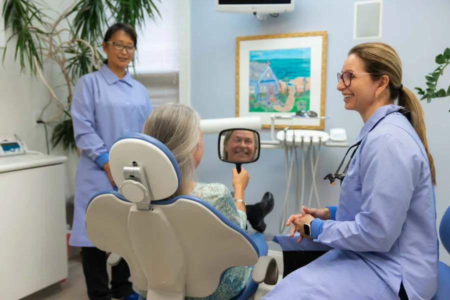 Dentist and assistant smiling as elderly patient checks her smile in handheld mirror in dental clinic.