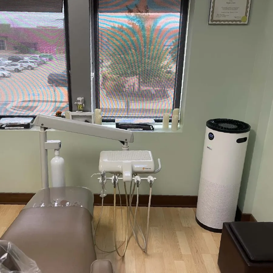 Modern dental office with a dental chair, equipment, air purifier, and two large windows showing a parking lot outside.