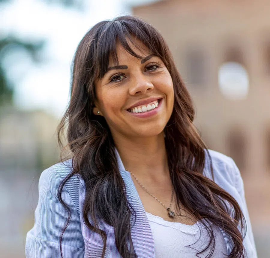 Smiling woman with long brown hair wearing a light blazer and white top outdoors in natural light.
